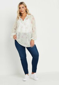 Blusa floreale bianca trasparente con scollo arricciato e coulisse, caratterizzata da fiori rosa pastello e pois verdi. Abbinata a jeans scuri e sneaker bianche.