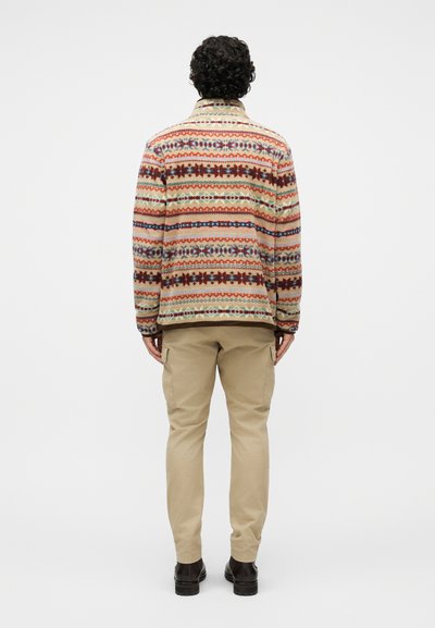 Mees, kes kannab beežikujulisi cargo-pükse, tumedaid jalatseid ja mitmevärvilist mustrilist fliisjopet, seisab näoga eemale ühevärvilise tausta ees.