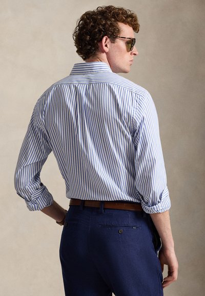 Striped blue and white shirt with rolled sleeves, collared neckline, and button placket. Paired with dark blue trousers and brown belt.