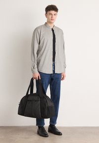 Svart duffelbag med teksturert stoff, langstrakt form og merke på fronten. Grå skjorte med ståkrage, paret med blå jeans og svarte sko.