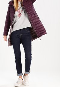 Chaqueta morada acolchada con cremallera, sudadera gris con estampado floral, jeans oscuros arremangados en los puños y zapatillas claras sobre un fondo neutro.