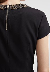 Schwarzes Kleid mit einem beliebten Kragen und metallischen Akzenten. Reißverschluss hinten, kurze Ärmel und strukturiertes Gewebe.