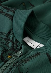 Zelená svetr s zipem a měkkými lemy na límci a manžetách s černým potiskem. Zip je z plastu ve stejné zelené barvě. Štítek uvnitř.