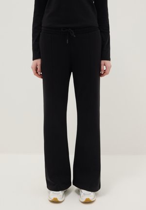 Mujer con pantalones negros de pierna ancha y cintura con cordón, y zapatillas blancas, posando contra un fondo claro y liso.