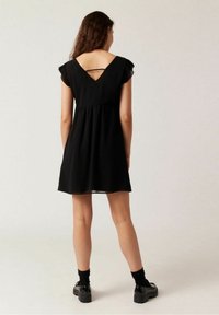 Svart klänning med korta volangärmar, en samlad midja och en nyckelhålsöppning på baksidan. Slätt tyg med en flödande silhuett.