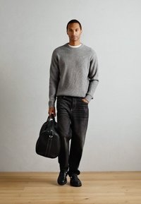 Gray ribbed sweater, dark jeans, black shoes, holding a black leather bag. Background shows a light gray wall and wooden floor.