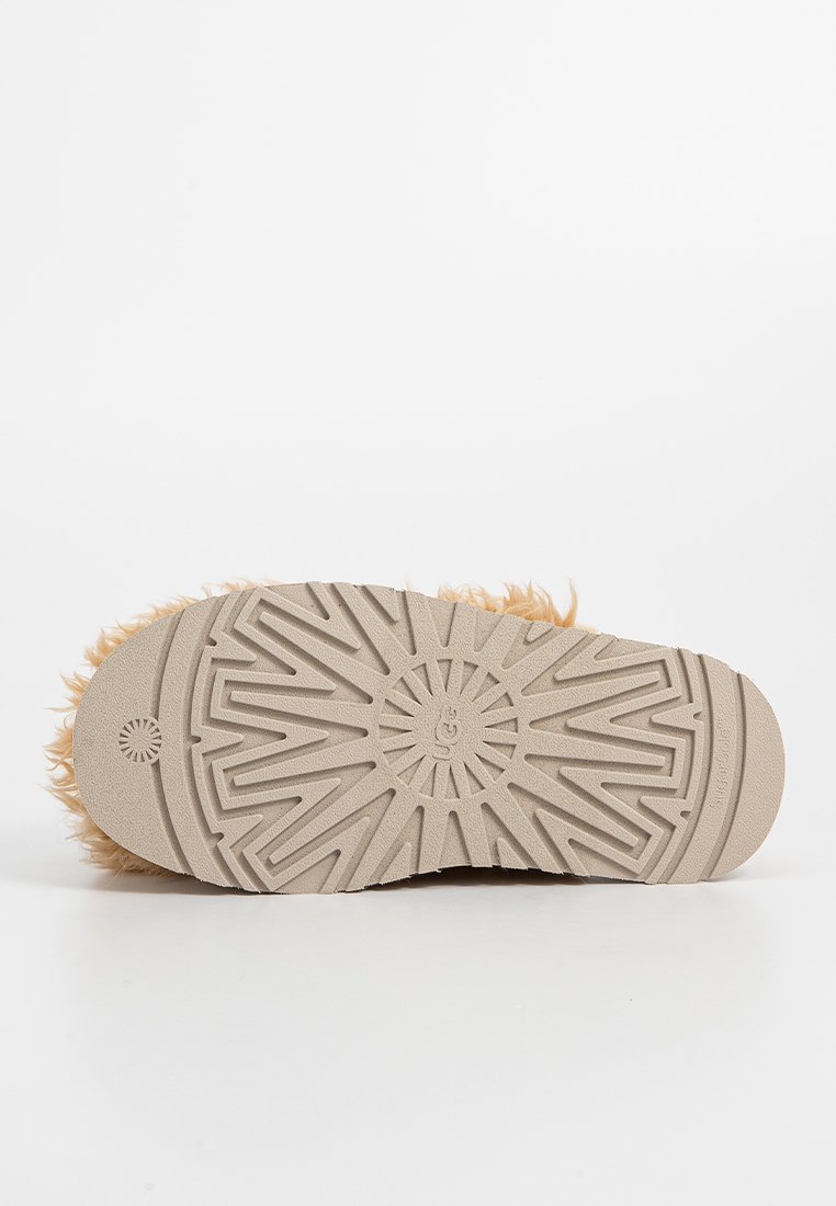 Semelle d'un chausson UGG beige avec un motif de semelle en zigzag et des bords en fourrure tan duveteuse visibles autour des côtés.