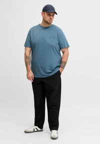 Blaues Kurzarm-T-Shirt mit kleinem Logo, kombiniert mit schwarzer Hose und weißen Sneakers mit schwarzen und grauen Akzenten.