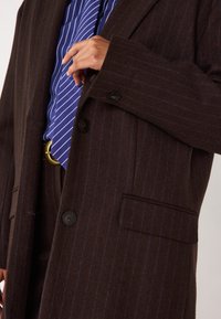 Cappotto marrone a righe fatto di lana, con bottoni neri, due tasche e stratificato con una camicia a righe blu e bianche sotto.