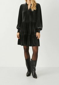 Robe en velours noir à manches longues, col en V, et jupe à étages. Portée avec des collants noirs et des bottes noires montantes jusqu'au genou. Texture lisse.