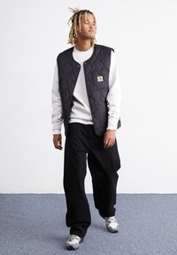 Quilted black vest over a grey sweatshirt, paired with loose black pants and grey sneakers. Features a small logo patch on the vest.