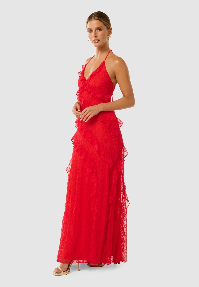Robe maxi en dentelle rouge avec des couches à volants, décolleté en V plongeant, bretelles halter, et silhouette fluide ; portée avec des talons dorés à lanières.