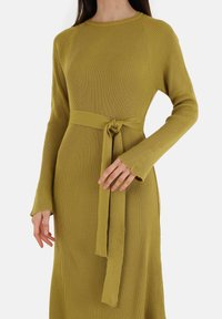 Vestito a maniche lunghe a coste di colore verde oliva, con vita sbloccata e dettaglio annodato, e scollatura a girocollo. Tessuto morbido e vestibilità affusolata.