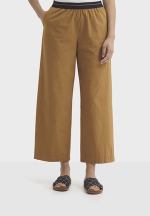 Personne portant un pantalon court à jambes larges couleur moutarde avec une taille élastique et des sandales slip-on noires à brides tressées.