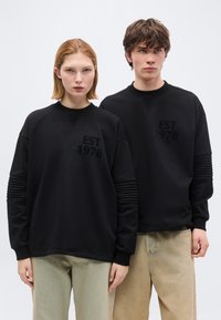 Δύο νεαροί ενήλικες φορούν μαύρα φούτερ με την επιγραφή "EST 1978", στέκονται δίπλα δίπλα μπροστά σε μια απλή λευκή επιφάνεια.