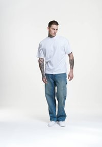 Camiseta blanca de manga corta combinada con pantalones vaqueros holgados de color azul claro con bolsillos tipo cargo, complementada con zapatillas blancas y tatuajes visibles.