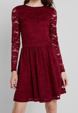 Robe en dentelle bordeaux avec manches longues transparentes, taille cintrée et jupe évasée portée par une femme debout sur un fond uni.