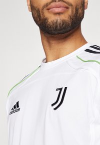 Biała koszulka sportowa z czarnym logo Adidas i herbem Juventusu. Zielone akcenty na ramionach, czarne paski na rękawach.