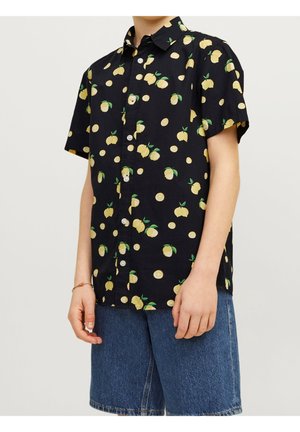 Person iført en sort kortærmet skjorte med gule frugtprint og blå denimshorts, stående mod en ensfarvet baggrund.