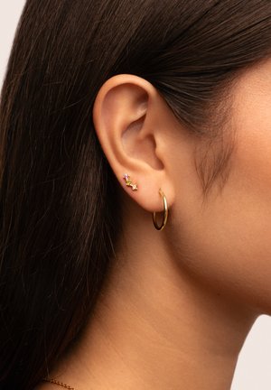 Goldene Creolenohrringe, ergänzt durch drei kleine Edelsteine in Pink und Gelb, die an dem Ohr eines Modells von der Seite präsentiert werden, um die Ohrmerkmale hervorzuheben.