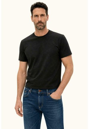 Mężczyzna z ciemnymi włosami i brodą, ubrany w czarny t-shirt z krótkim rękawem i niebieskie dżinsy, z jedną ręką w kieszeni, na neutralnym tle.
