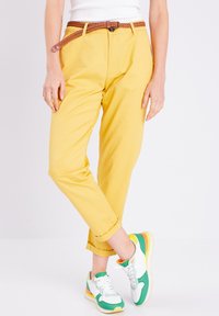 Pantalons en coton jaunes avec une ceinture tressée marron ; associés à des baskets multicolores avec des accents verts, blancs et jaunes.