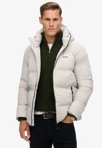 Lichte grijze pufferjack met capuchon, voorzien van een rits aan de voorkant, zwarte accenten en een gestructureerde groene gebreide trui eronder.