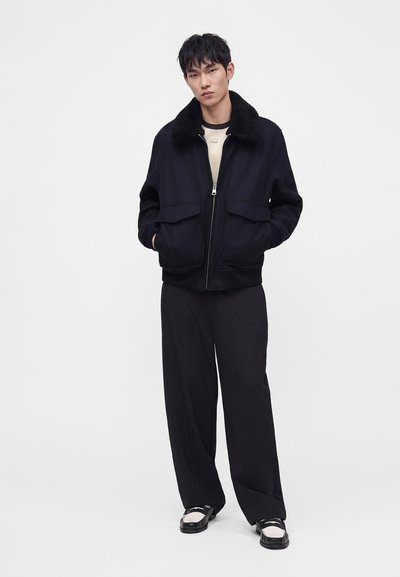 Modèle masculin portant une veste bomber noire avec un col en fourrure, un pantalon noir ample, une chemise blanche et des chaussures noires et blanches, debout devant un fond blanc.