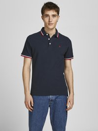 Mornarska polo majica s crvenim i bijelim obrubom oko ovratnika, dvobutonim kopčanjem i malim izvezenim logotipom na prsima; u kombinaciji s plavim farmerkama.