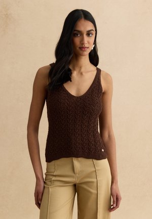 Donna con lunghi capelli neri che indossa un top in maglia senza maniche marrone scuro e pantaloni beige a vita alta, in piedi su uno sfondo beige.