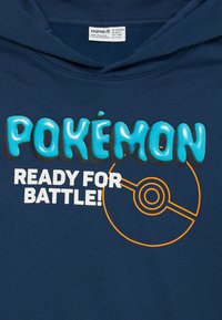 Tumši zils kokvilnas kapucis, ar uzrakstu "POKÉMON" turkoza krāsā un "READY FOR BATTLE!" baltā krāsā, papildināts ar oranžu Poké Ball grafiku.