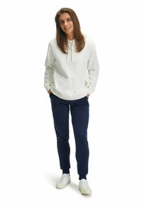 Witte hoodie met een koord en voorzakken, gecombineerd met marineblauwe joggingbroek. Model draagt witte sneakers. Eenvoudig, casual ontwerp.