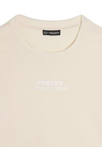 T-shirt in cotone color crema con scollo rotondo, caratterizzato da testo ricamato bianco: "FREDDY OGGI È IL TUO GIORNO." Texture liscia.