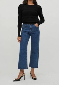 Zwarte gebreide trui met gestructureerde strepen, gecombineerd met lichtblauwe high-waisted broek met wijde pijpen en zwarte puntige hakken met gouden accenten.