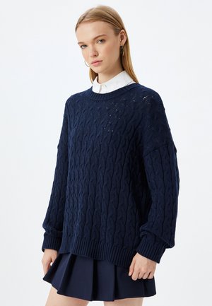 Maglione navy a trecce con scollatura rotonda, spalle scese e polsini a coste, abbinato a una gonna navy plissettata.