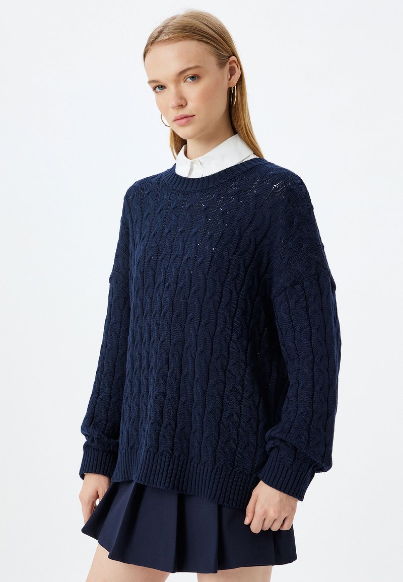 Granatowy sweter w splot kabelkowy z okrągłym dekoltem, opuszczonymi ramionami i ściągaczami na mankietach, zestawiony z plisowaną granatową spódnicą.