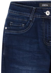 Jean en denim bleu foncé avec poches avant, bouton argenté, passants de ceinture et surpiqûres contrastées, étiquette de marque "CECIL" à l'intérieur de la ceinture.