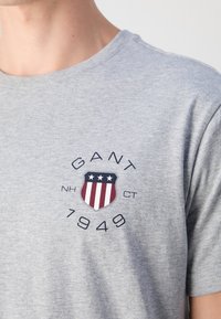 Gros plan sur un t-shirt gris avec un logo en forme de bouclier comportant des étoiles et des rayures, entouré des textes « GANT », « NH », « CT » et « 1949 ».