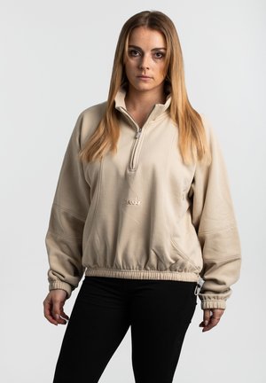 Beiger Fleecepullover mit hohem Kragen, Halb-Reißverschluss, elastischem Bund und gerippten Bündchen. Verfügt über ein dezentes Logo auf der Vorderseite.