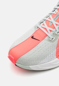 Grijze sportschoen met een koraalkleurige accent, lichte mesh bovenkant met perforaties en een witte zool. Opvallend Nike-logo in het zwart.