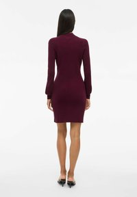 Robe en tricot côtelé bordeaux avec des manches longues et une silhouette ajustée, se terminant au milieu de la cuisse. Texture douce et design minimaliste.