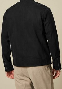 Schwarze Wildlederjacke mit einfachem Design, mit Reißverschluss versehenen Ärmelbündchen und glatter Textur; von hinten betrachtet. Taillierte Passform kombiniert mit beigen Hosen.