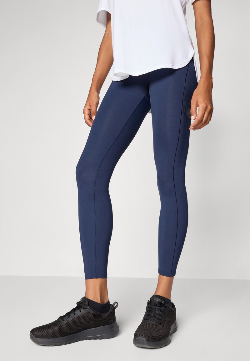 Eine Person steht in einem lockeren weißen Hemd, marineblauen Leggings und schwarzen Sport-Schuhen da und zeigt legere Sportbekleidung.