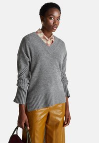 Šedý svetr s výstřihem do V a žebrovanými manžetami a lemem, vrstvený přes kostkovanou košili. Model nosí kožené hořčicové kalhoty a drží zelenou kabelku.