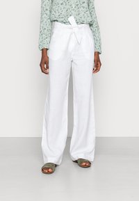 Person som bär högmidjade vita linnebyxor med vida ben och knytbälte, tillsammans med en grön blommig långärmad topp och olivgröna sandaler.