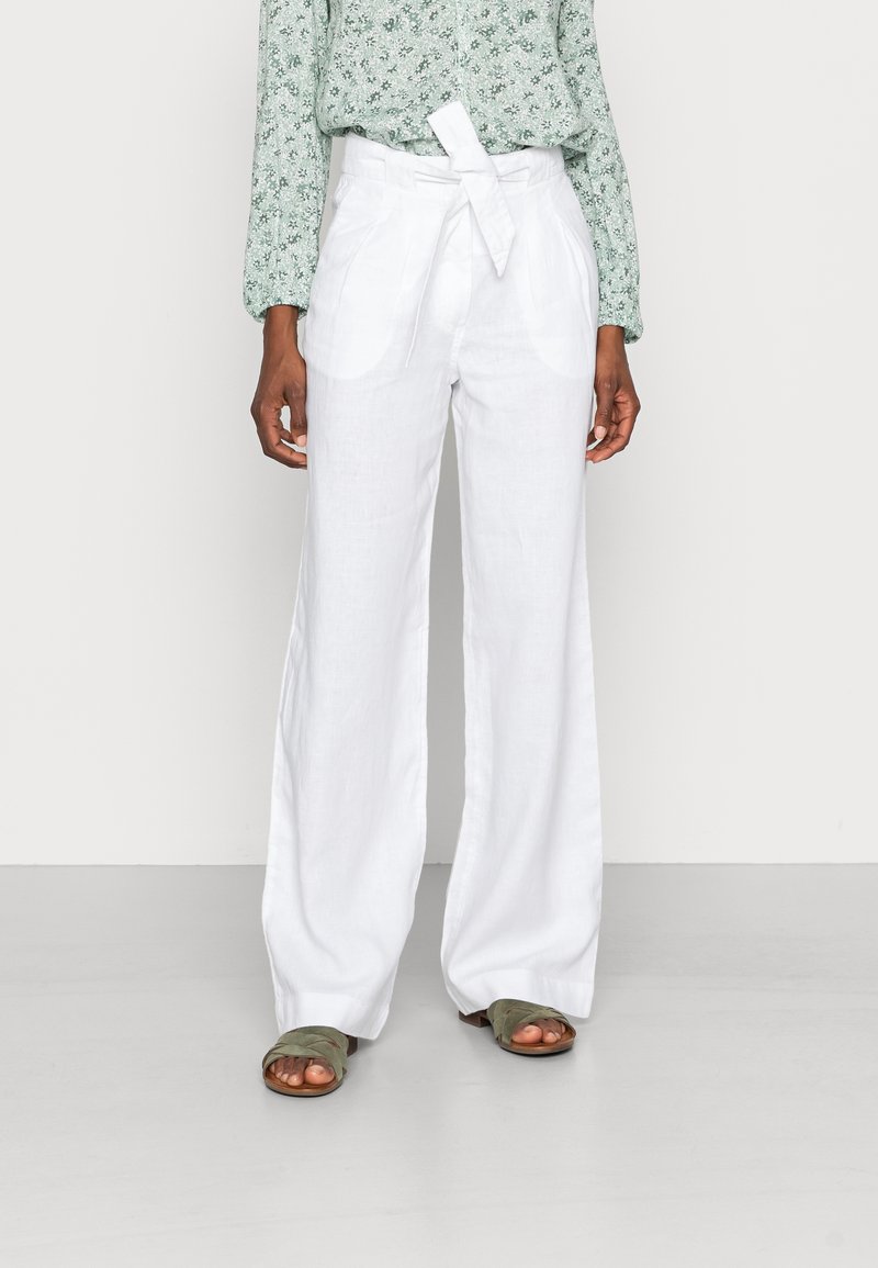 Person som bär högmidjade vita linnebyxor med vida ben och knytbälte, tillsammans med en grön blommig långärmad topp och olivgröna sandaler.