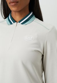 Close-up van een persoon in een beige langemouwen polo met ritssluiting en groene gestreepte afwerking, met het EA7 Emporio Armani-logo op de borst.