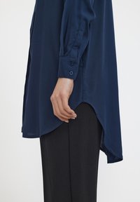 Chemise bleu marine à manches longues avec des boutons aux poignets, fentes sur les côtés et ourlet asymétrique, associée à un pantalon noir. Texture de tissu lisse.