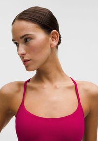 Žena s tmavými vlasmi zviazanými dozadu, oblečená v jasne ružovom bezrukávovom topu, pozerajúca doľava na pozadí s jednofarebným svetlým pozadím.