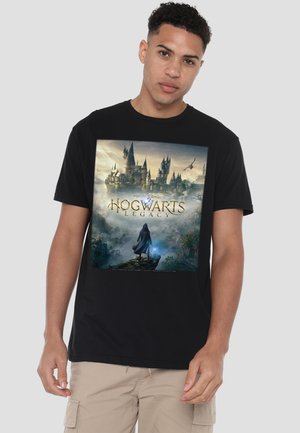 Young man wearing black t-shirt with Hogwarts castle and "Hogwarts Legacy" text, standing in misty magical landscape holding a glowing wand.